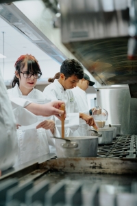 Students stirring food in a pot on a stove.