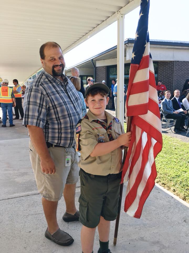 A smailing man stands behind a young person in a scout uniform holding an American flag