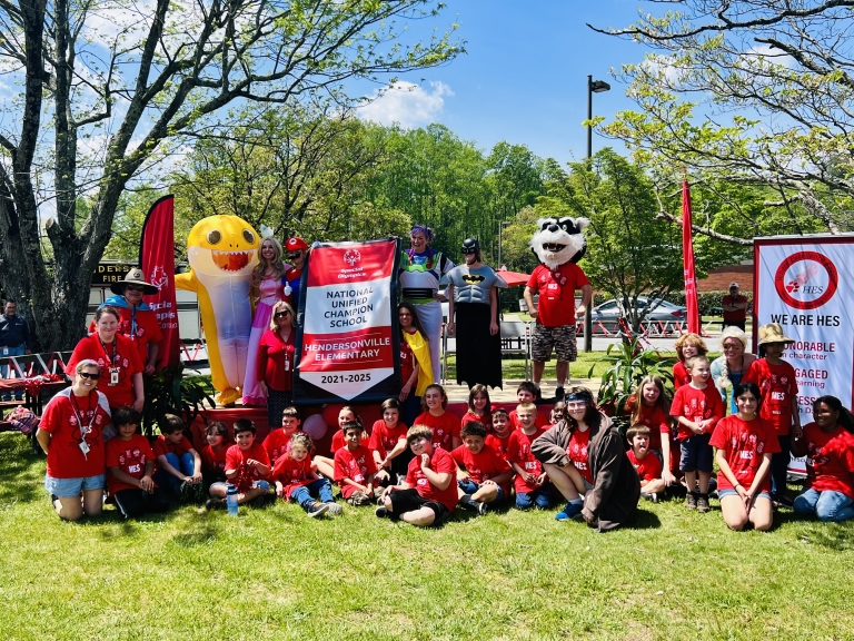 Group of students and staff members sitting and kneeling in front of a stage outdoors. Adults dressed as mascots and superheroes are on the stage behind them. Two adults are holding a banner that reads National Unified Champion School
