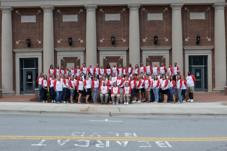 School staff in matching red and white shirts, stand on the sidewalk. The street in front of them has the words Bearcat Blvd painted on it
