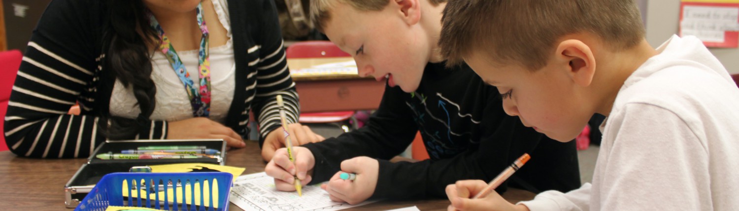 A teacher and two young boys sit at a table. the boys are coloiring while the teacher watches.