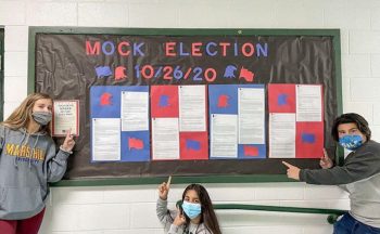 three high school students pointing to bulletin board with "Mock Election" on it