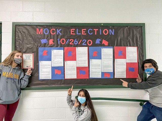 three high school students pointing to bulletin board with "Mock Election" on it