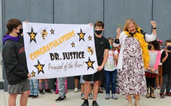 two boys in masks hold sign that says "congratulations dr. justice principal of the year" while woman does happy dance