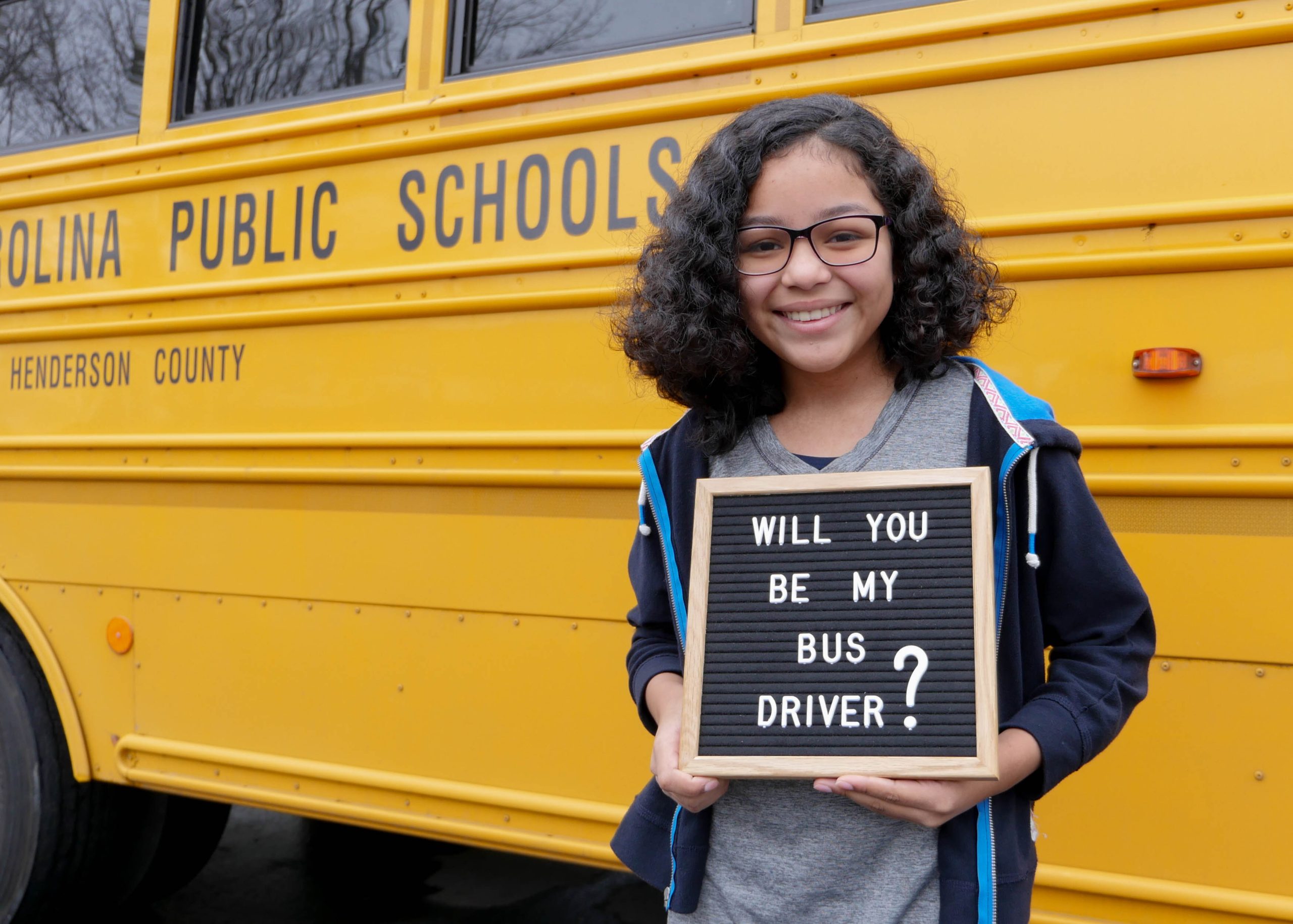 Student stands beside school bus holding sign that reads Will you be my bus driver?