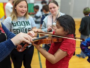 2 older students in background smile as younger student tries out violin