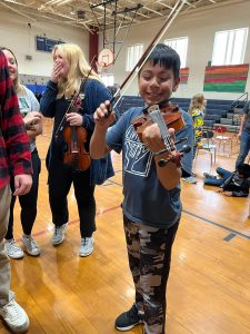 boy holding violin and smiling, 2 older students in background