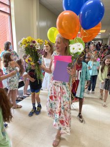 woman in hallway holding balloons and being handed flowers by students