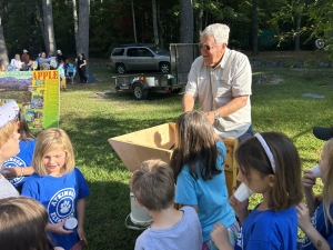 Students watching a presenter put apples in a apple press for apple cider. There are two students drinking the cider and four watching. 