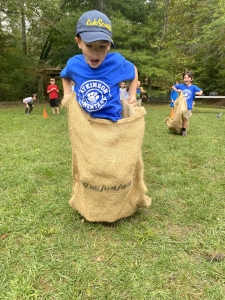 A student doing a sack race. In the background there are two students watching and two students also doing a sack race. 