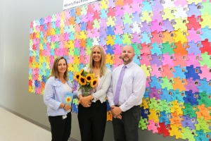 Ms. Schneider with Principal Andrew VunCannon and Vice Principal Ms. Rudi in front of a colorful puzzle piece wall. 