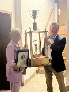 Sue Moon holds a plaque and big trophy with another man at the other side of the trophy. 