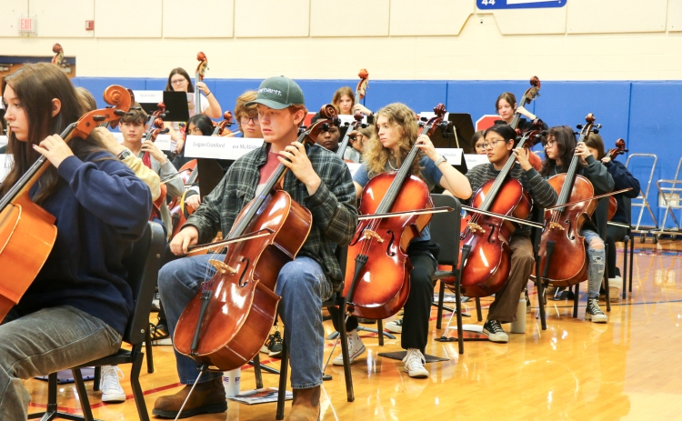 An image of over a dozen orchestra students sitting in chairs during rehearsal practice in the gym