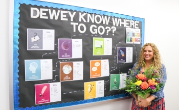 Ms. Cheek holding flowers in front of a Dewey Decimal System bulletin board