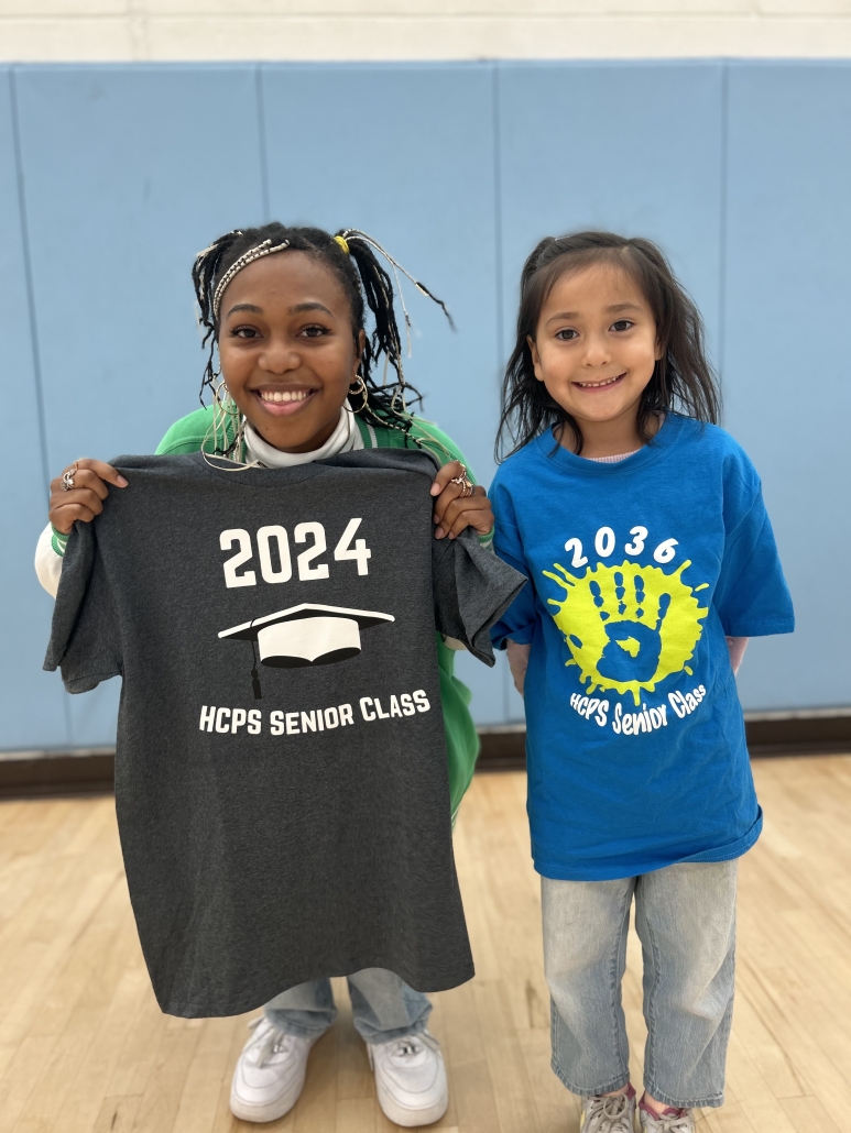 A senior holding a class of 2024 shirt while standing next to a kindergartener wearing a class of 2036 shirt. 