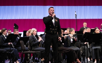 A photo of a man in uniform singing on stage with over a dozen band members also in uniform.