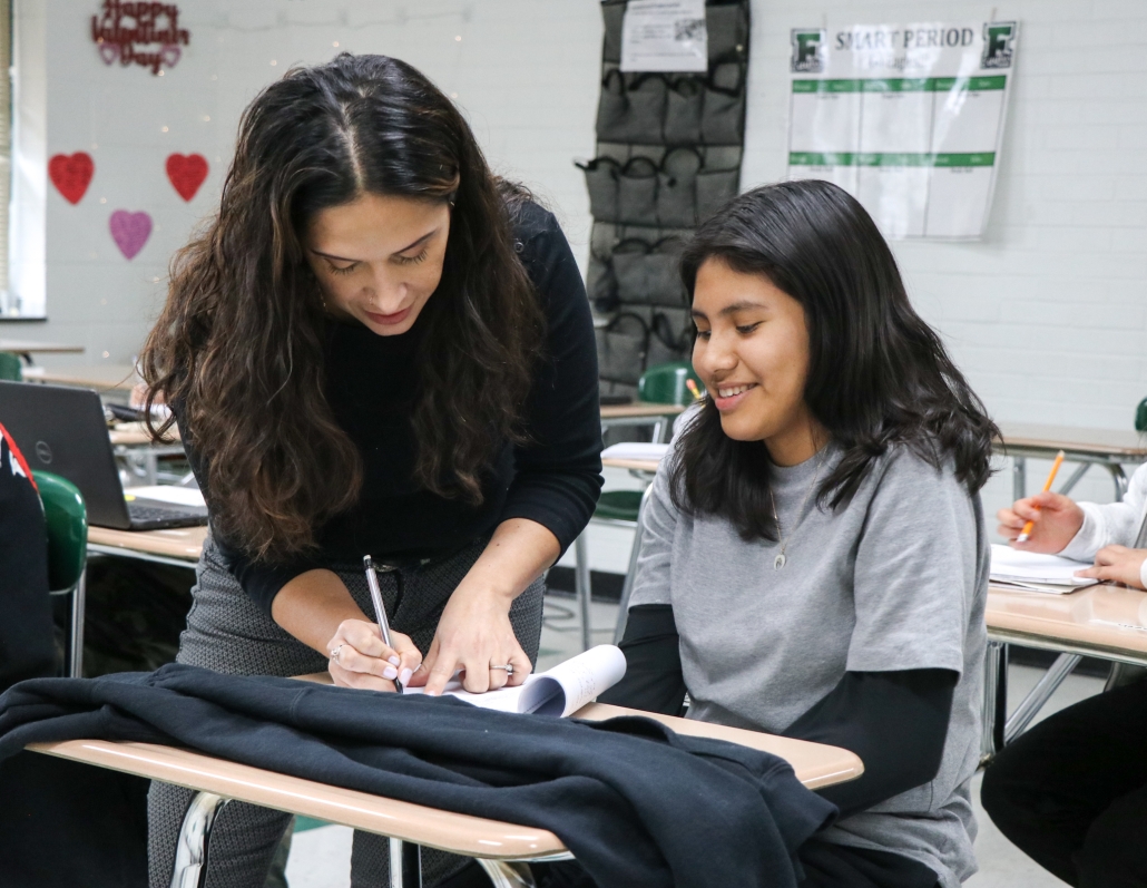 Johanna Bosch-Diaz leaning over a desk writing while helping a female student. The student is smiling in the desk. 