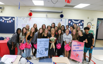 Mrs. Stepp surrounded by 22 students in her classroom. Everyone is posing for a picture and there are several pink pom poms and signs in the photo.
