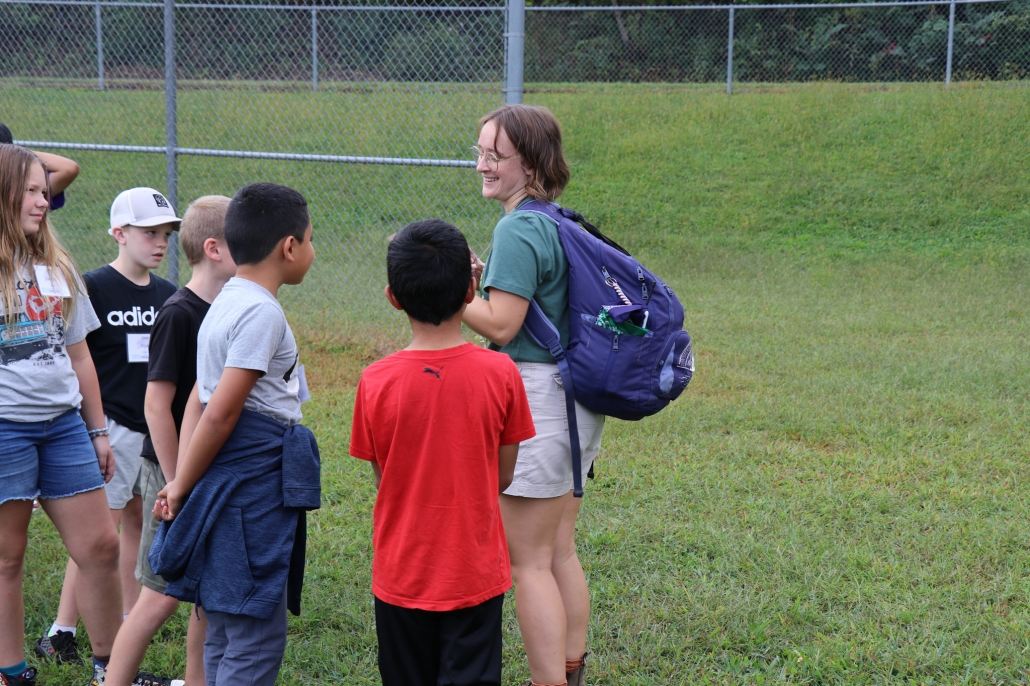 A mountain roots instructor with a backpack on talking with five students outside.