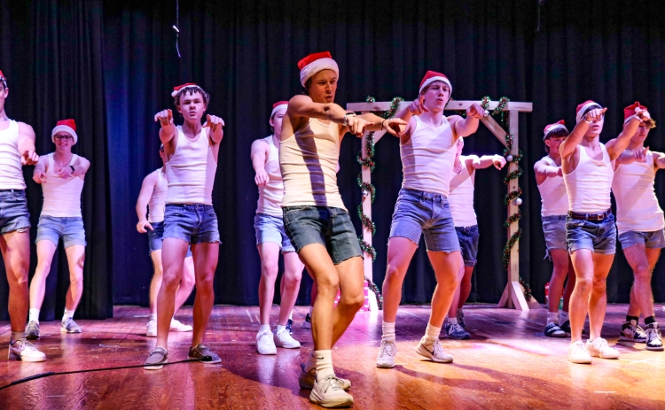 About 10 Mr. WHHS contestants on the stage in matching white tank tops and blue jean shorts. They are dancing with santa hats on.
