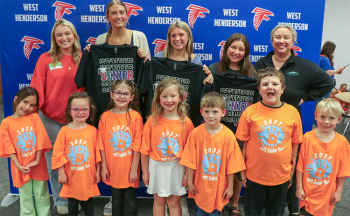 Three seniors hold up their senior t-shirts while they pose for a photo with seven kindergarteners and two sponsors. They are taking the photo in front of a West henderson falcon backdrop. The kindergarteners are wearing orange Class of 2037 t-shirts.