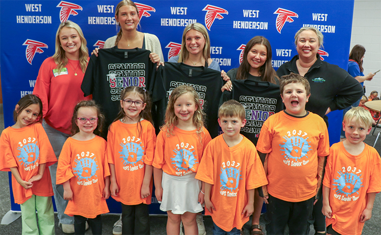 Three seniors hold up their senior t-shirts while they pose for a photo with seven kindergarteners and two sponsors. They are taking the photo in front of a West henderson falcon backdrop. The kindergarteners are wearing orange Class of 2037 t-shirts.