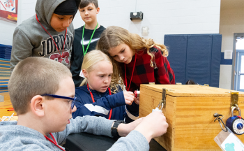 A group of five elementary school students collaborate intently to unlock a large wooden box with combination locks, participating in a classroom or gymnasium-based problem-solving activity.
