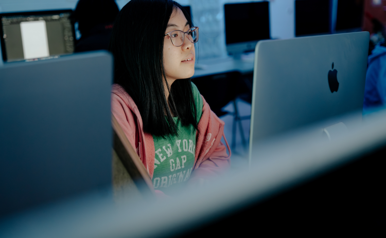A student with long dark hair and glasses works on a computer in a classroom.