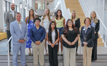 Students and representatives from HCPS, Blue Ridge, AdventHealth, and Pardee pose for a group photo during the Healthcare Fellows Signing Day celebration. The event takes place inside a bright, modern building with a wooden staircase and large windows in the background. Students stand proudly in the front and middle rows, with administrators and staff standing behind them in support.