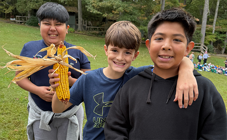 Three boys standing together and one is holding a dried corn husk. They are all three smiling.