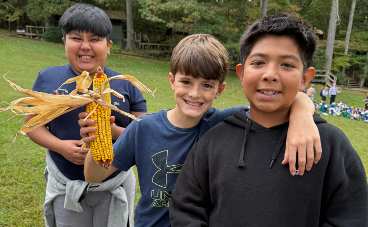 Three boys standing together and one is holding a dried corn husk. They are all three smiling.