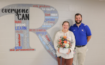 Molly Wright standing with Dr. John McDaris in front of a Rugby "R" mural in the hallway. Molly is holding flowers.