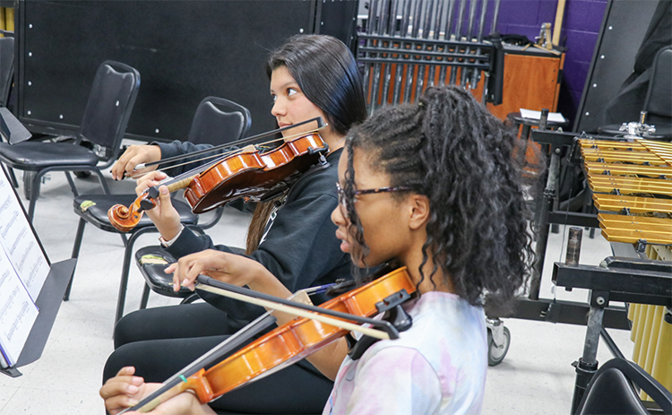 Two students playing violin while sitting in chairs in a music room during rehearsal.